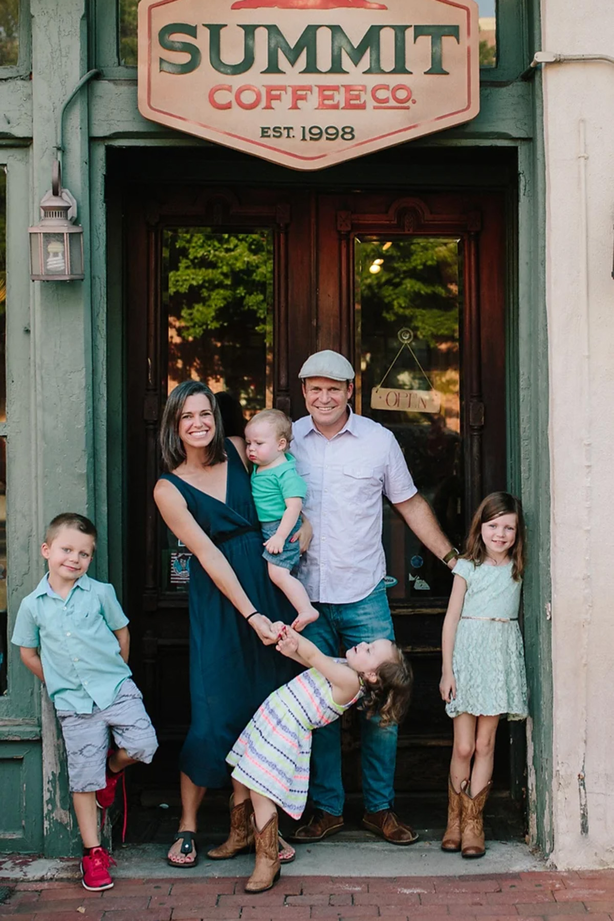 Beth Helfrich, a mother of five, stands outside a coffee shop with her family. She smiles while holding her youngest child, with her husband standing next to her, and her other children surrounding them.