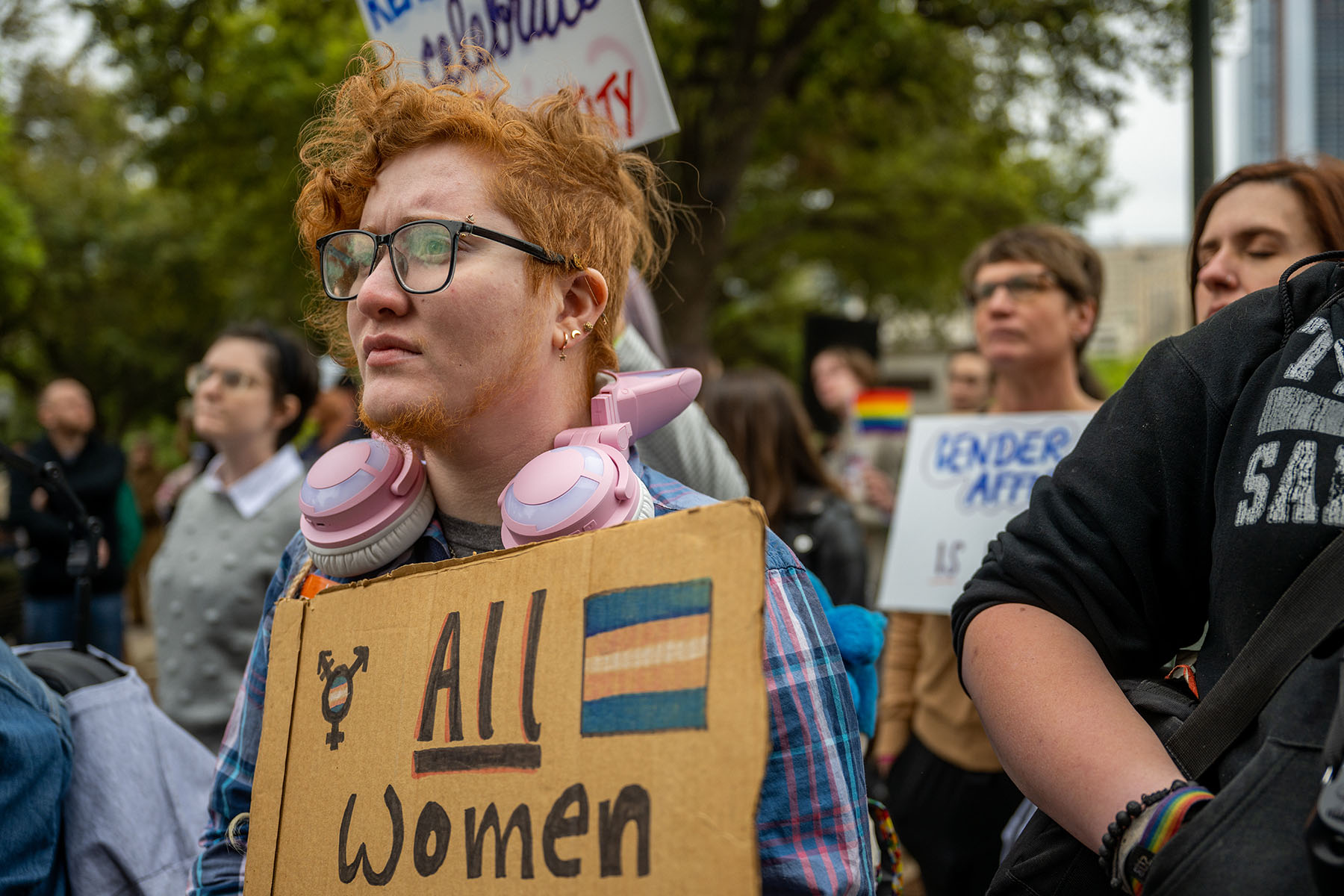 A young person with red hair and glasses, wearing pink headphones around their neck, holds a cardboard sign that reads "All Women" with a transgender symbol, while listening during a protest against anti-trans and LGBTQ+ legislation in Austin, Texas