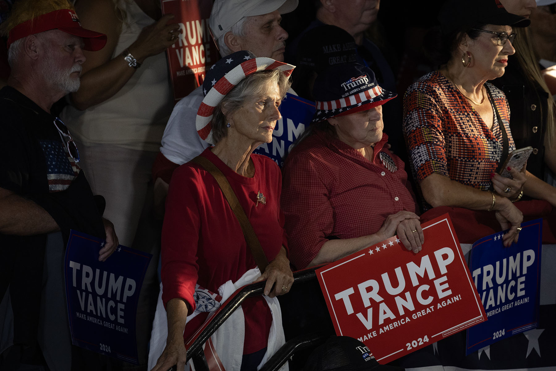 A group of women listen as Sen. JD Vance speaks at a campaign event, some wearing patriotic hats adorned with American flag designs, holding signs that read 'TRUMP VANCE 2024.'