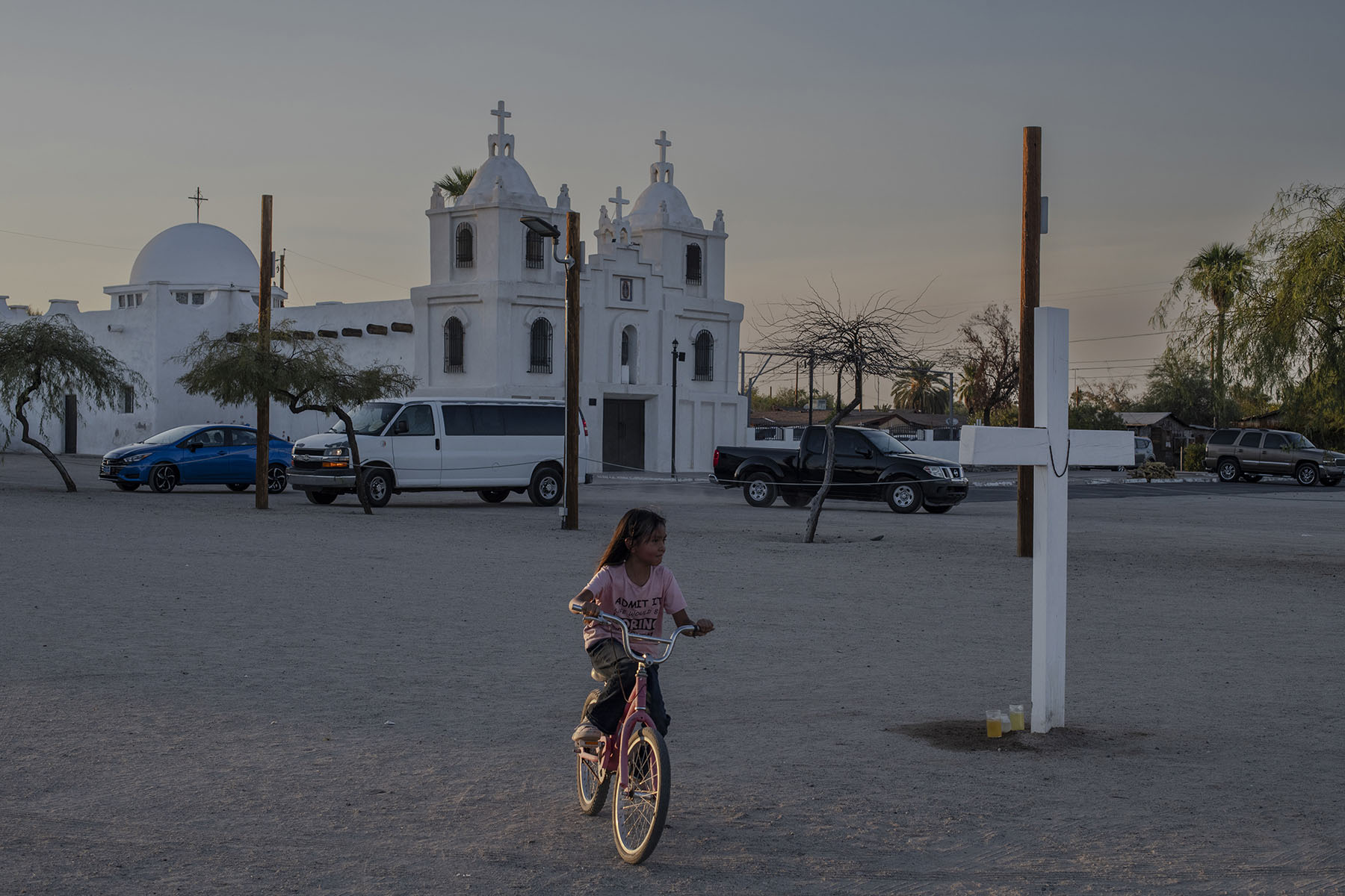 A young girl rides a bicycle in an open square near a white church with two bell towers. The scene includes parked cars and a large cross in the foreground.