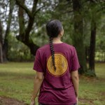 A native woman stands in a forest with her back facing the camera in this anonymized portrait.