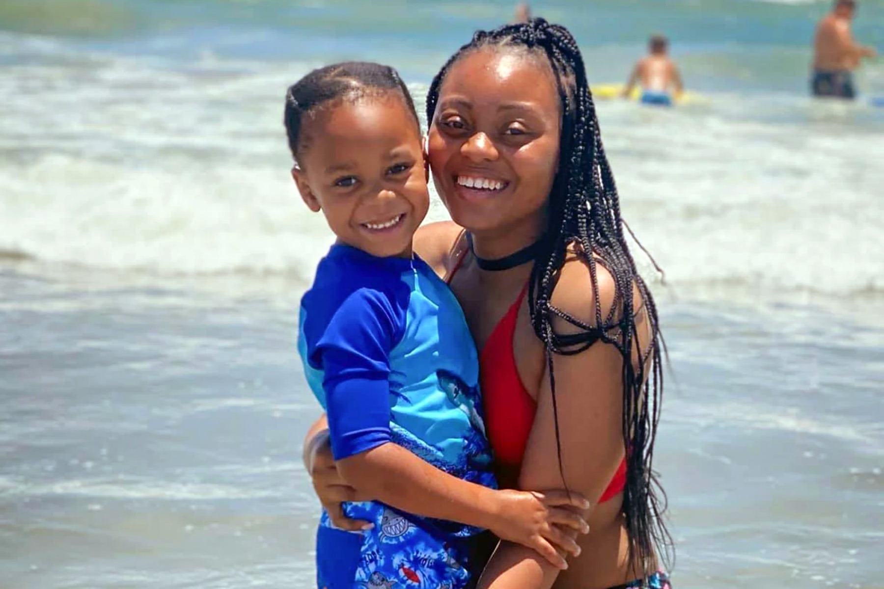 A smiling Amber Nicole Thurman in a red swimsuit holding her young son at the beach, standing close together in the water, with waves and other beachgoers in the background.