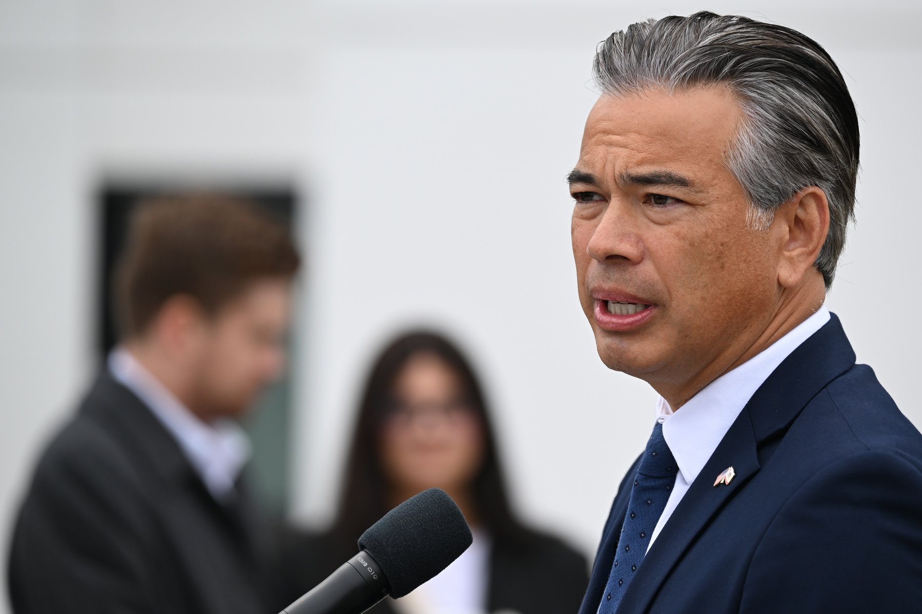 Attorney General Rob Bonta stands at a podium speaking with people in the background.