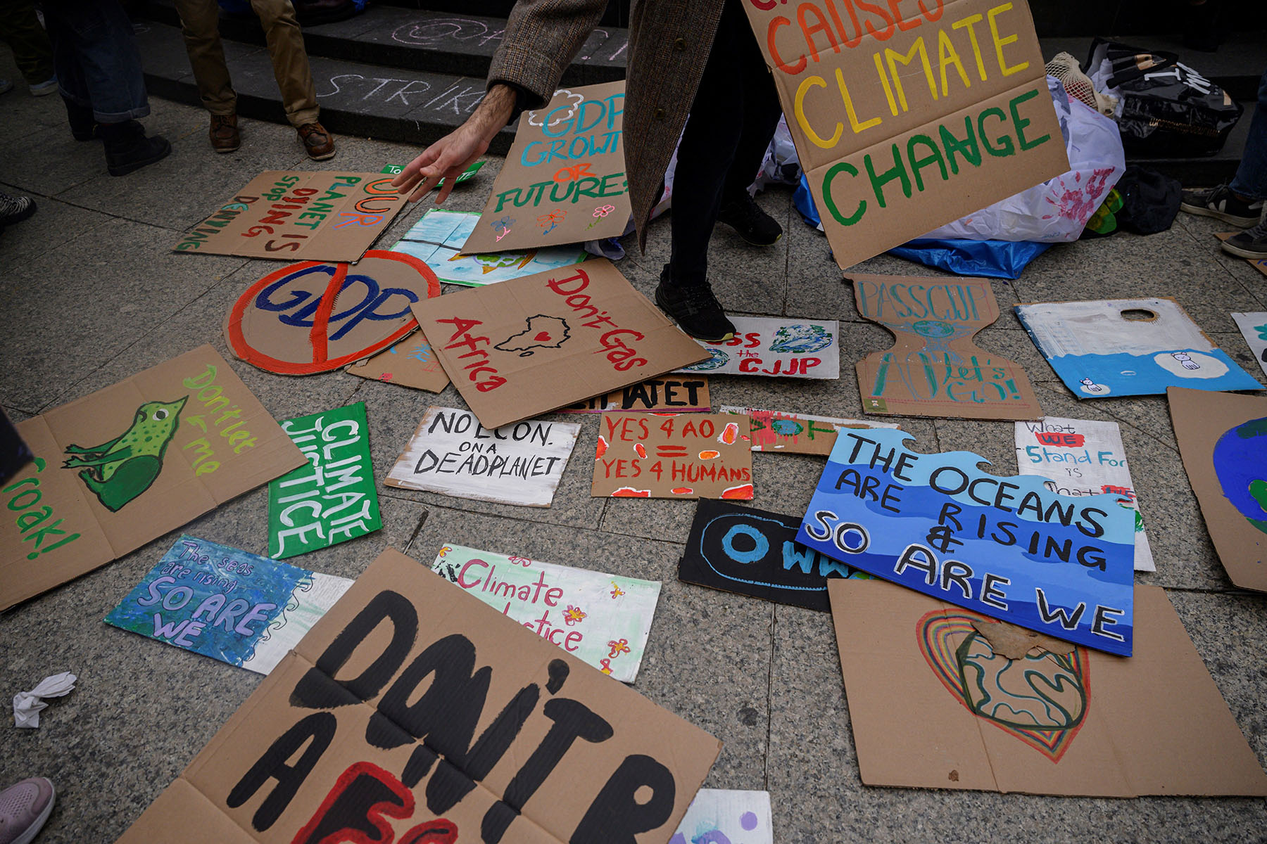 A person reaches down to pick up colorful protest placards scattered on the ground, with messages advocating for climate justice and against fossil fuels.