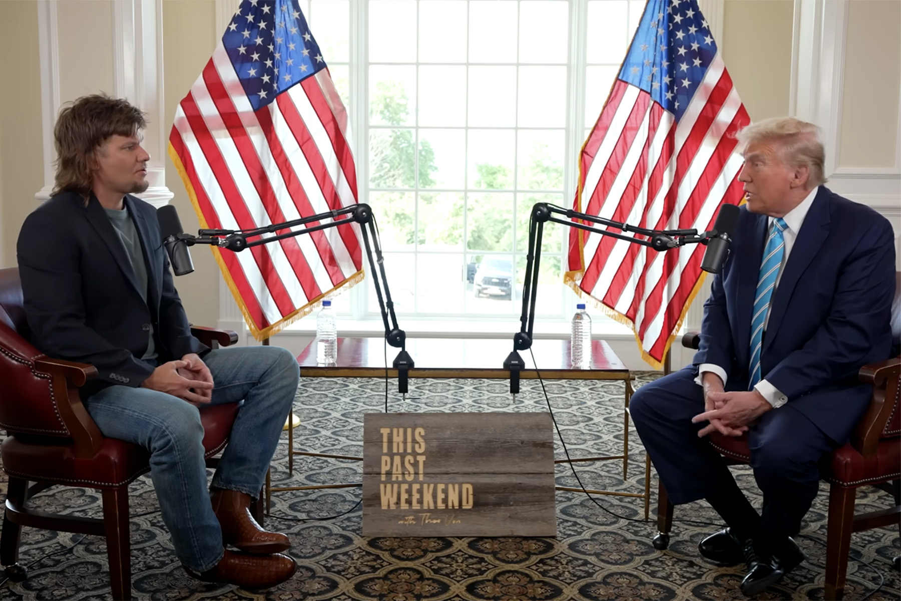 Former President Donald Trump and comedian Theo Von during an interview on "This Past Weekend," with both men seated in a room adorned with two American flags and microphone setups.