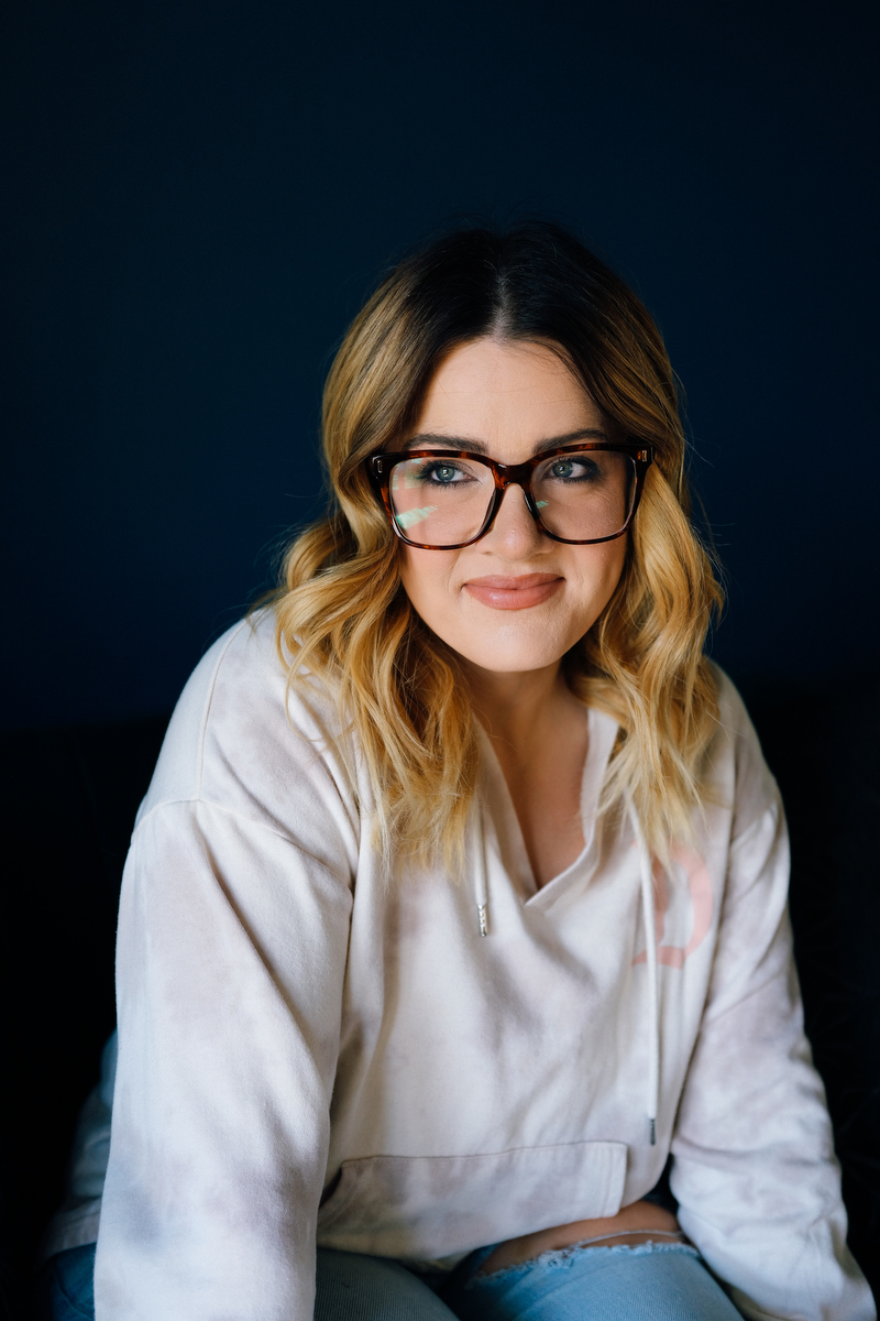 A portrait of a woman wearing glasses against a dark blue wall.