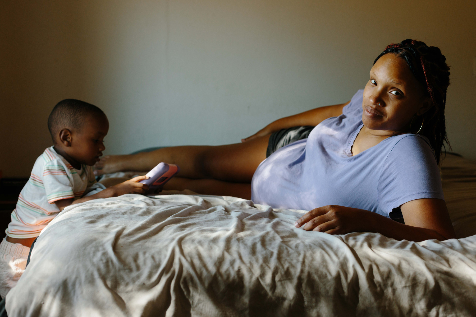 A pregnant woman lays across a bed while a toddler plays near her.