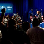 Vice President Kamala Harris speaking at a podium with American flags behind her, addressing a crowd at a campaign rally in Atlanta. A large sign behind her reads, '1 in 3 women lives under a Trump abortion ban.'