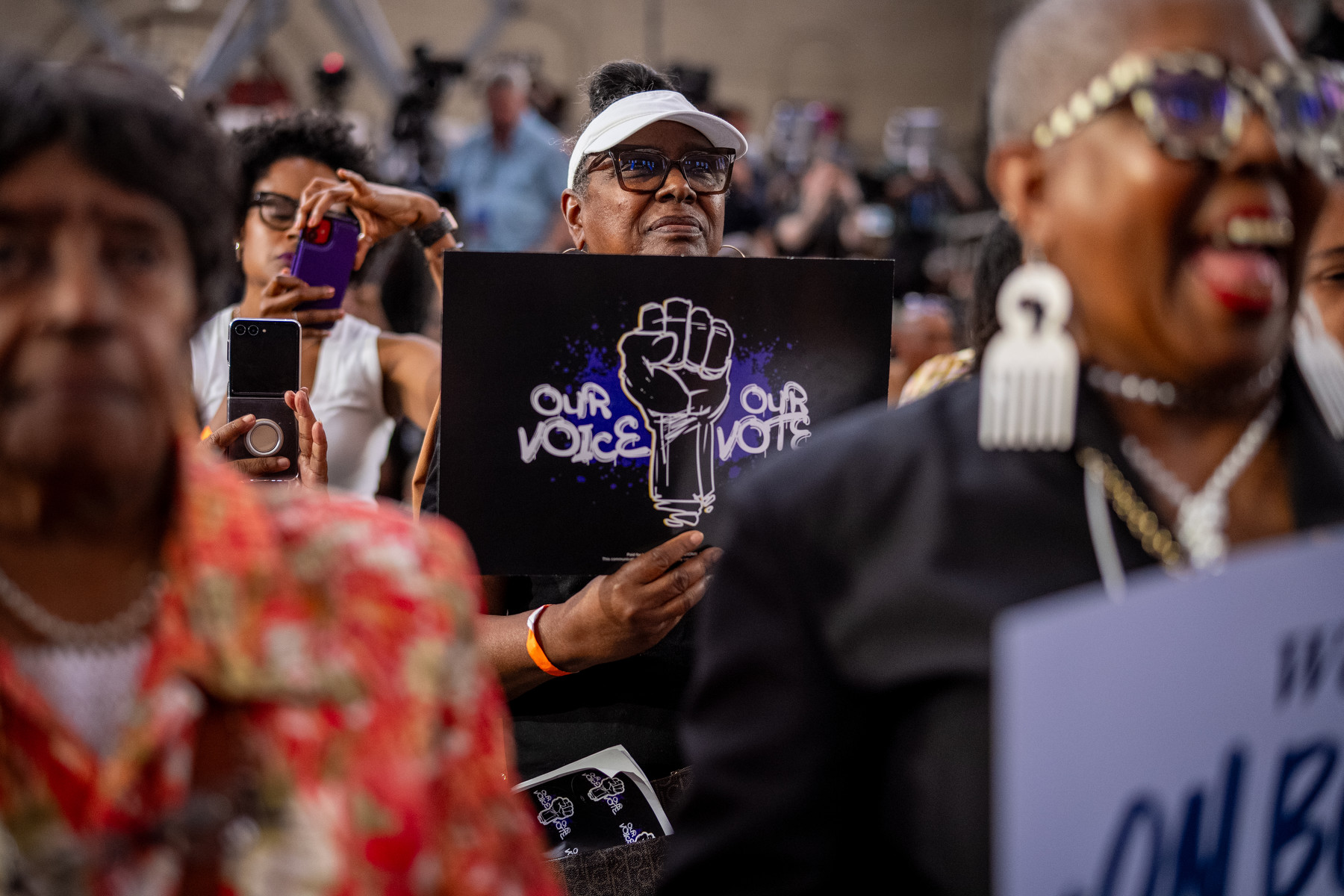 A crowd of Black women laugh and hold signs saying "our voice, our vote" at a campaign rally.