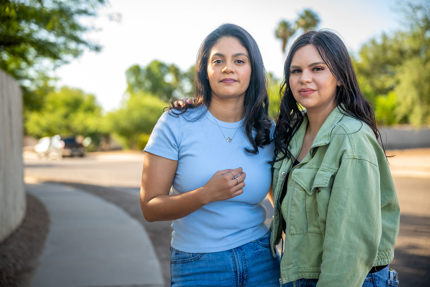 Adriana Grijalva and her sister stand close together, posing for a portrait at their home in Tucson, Arizona. The two women look at the camera, with a suburban street and greenery visible in the background.