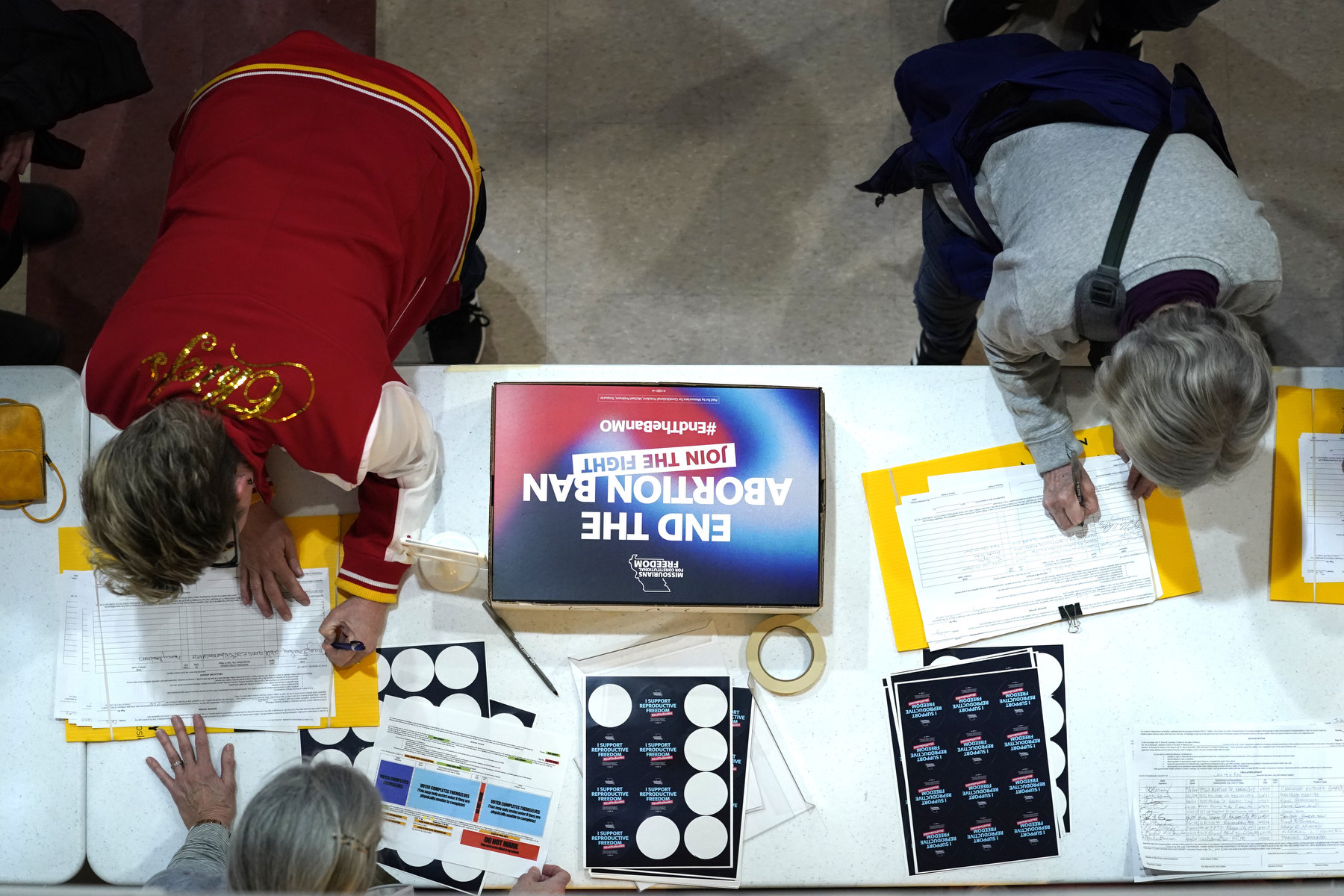 Two people sign petitions on a table, viewed from above.