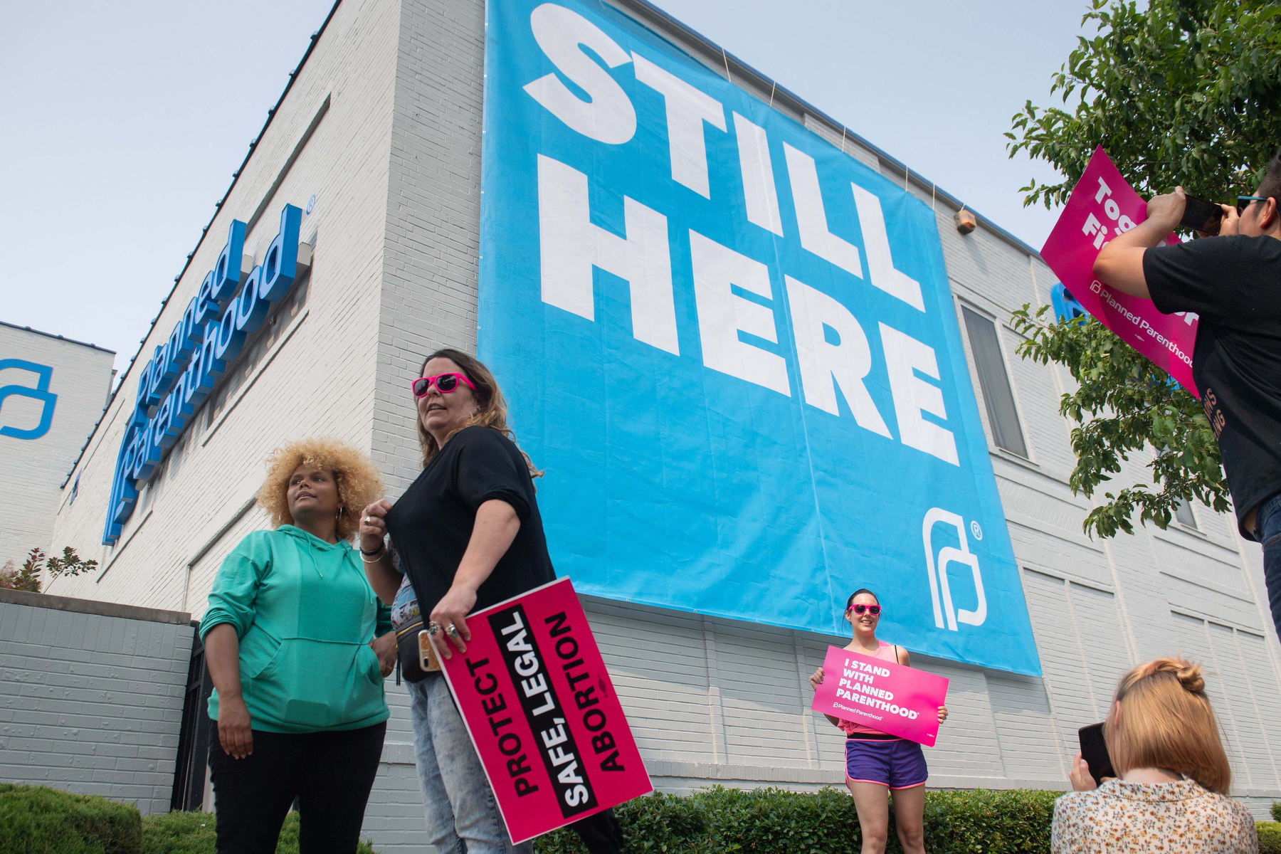 People stand around the outside of a Planned Parenthood building, taking photos in front of a sign that says "still here"