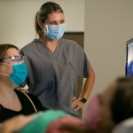 Dr. Kirstin Lyerly in medical scrubs stands beside another medical professional during a patient consultation, both wearing masks.