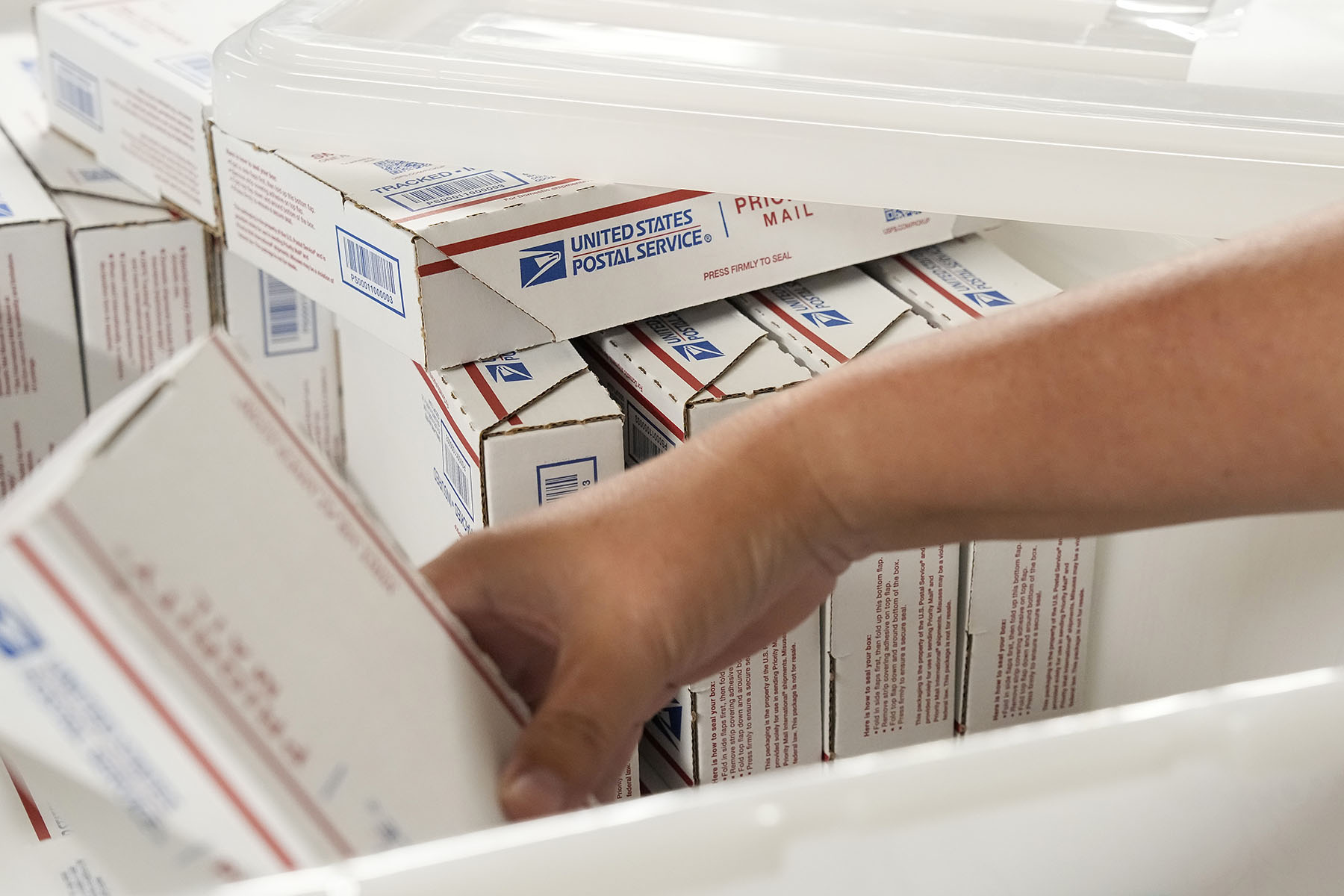 A close-up of a hand reaching for a box among many stacked United States Postal Service packages containing abortion medication.