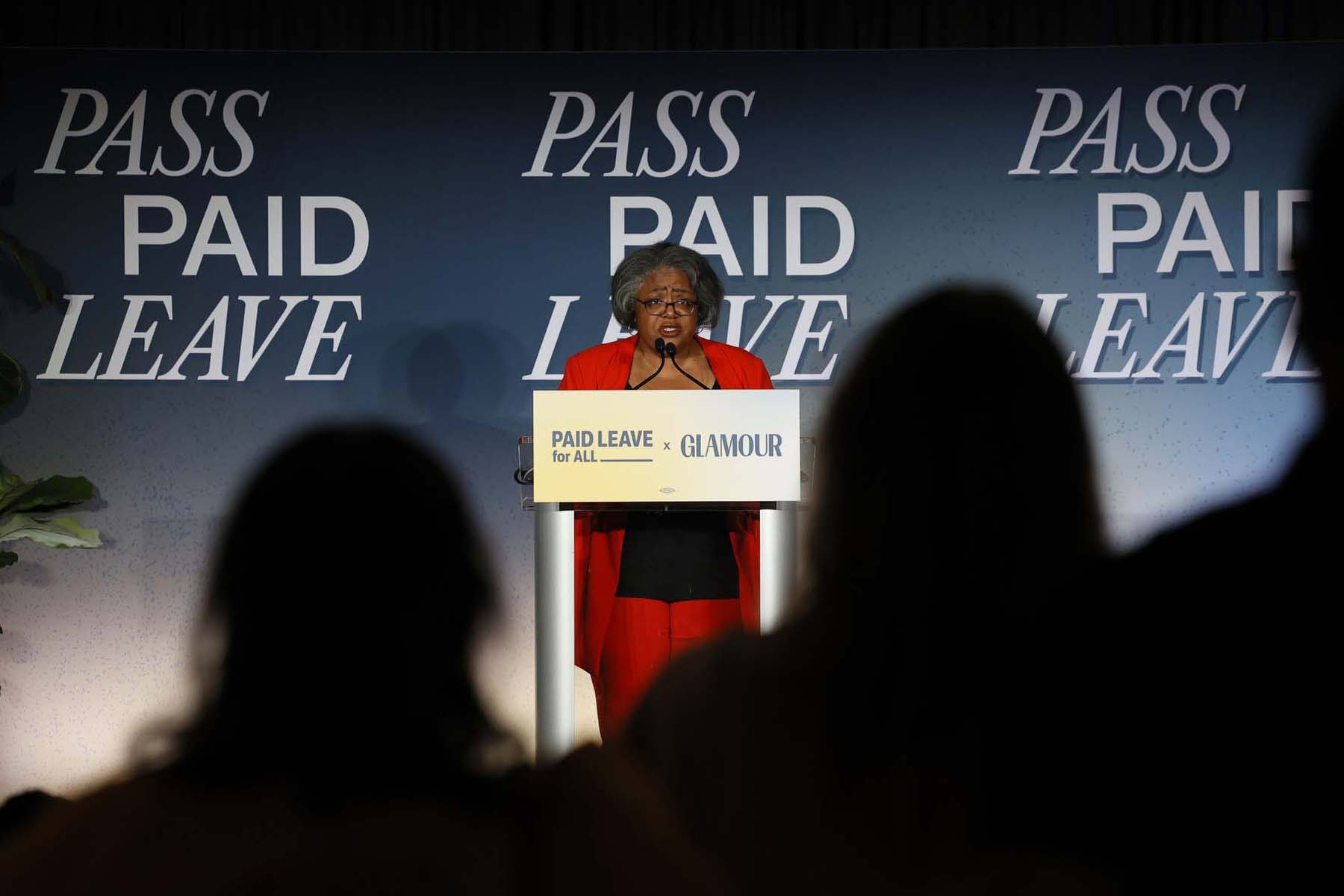 A woman stands at a podium with silhouetted audience members in the foreground.