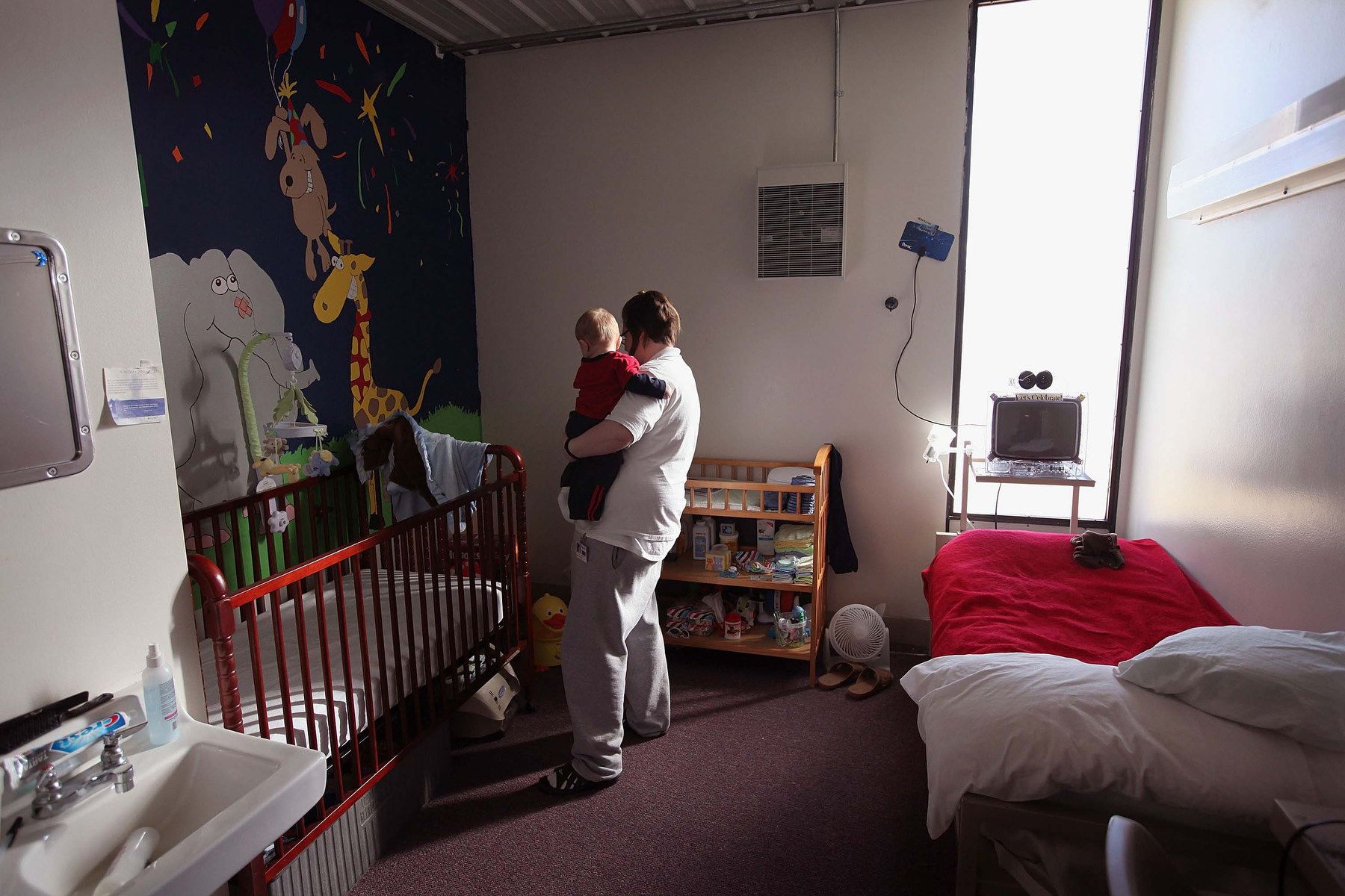 A mother holds an infant near a crib in a prison cell with a cartoon scene painted on the wall behind the crib.