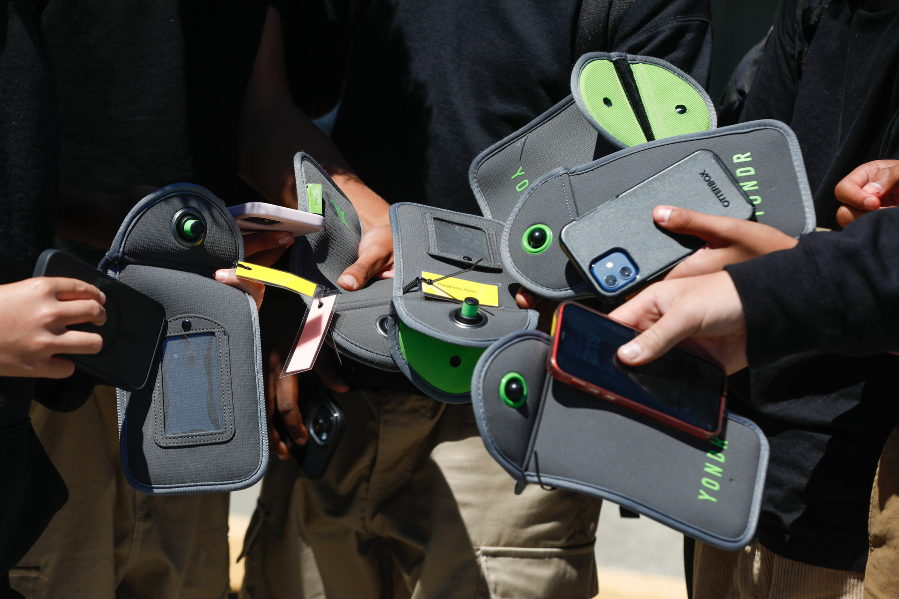 Several young hands hold flat gray pouches alongside cell phones as a group of students stand together.