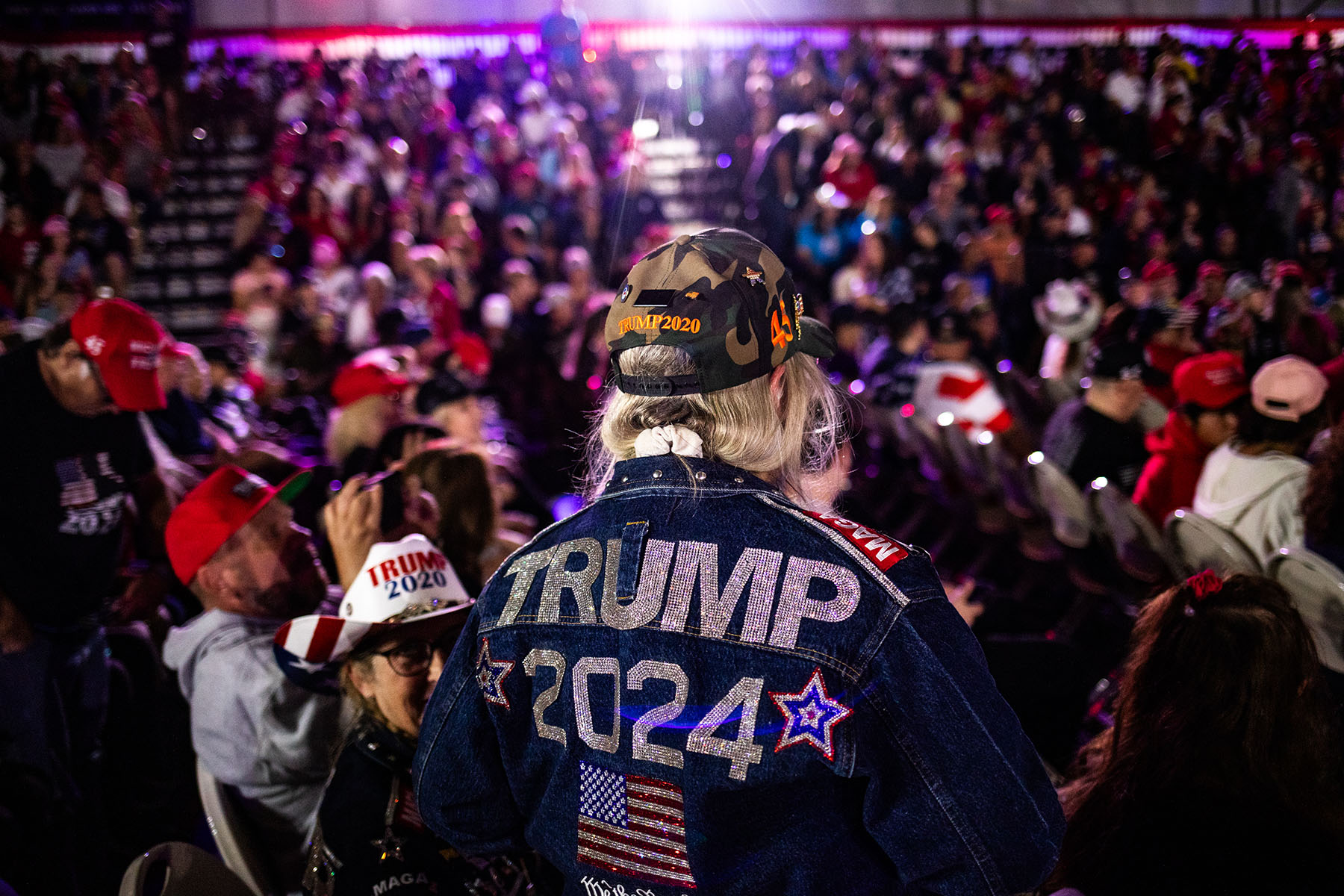 A crowd of people attend a Town Hall event with former President Trump, with a focus on a woman wearing a denim jacket with "Trump 2024" emblazoned in large letters on the back, along with patriotic symbols and an American flag.