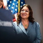 Lori Chavez-DeRemer stands in a blue suit and glasses in front of an American flag, preparing to speak at an event.