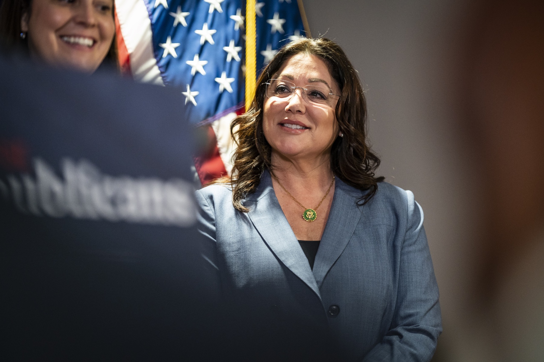 Lori Chavez-DeRemer stands in a blue suit and glasses in front of an American flag, preparing to speak at an event.