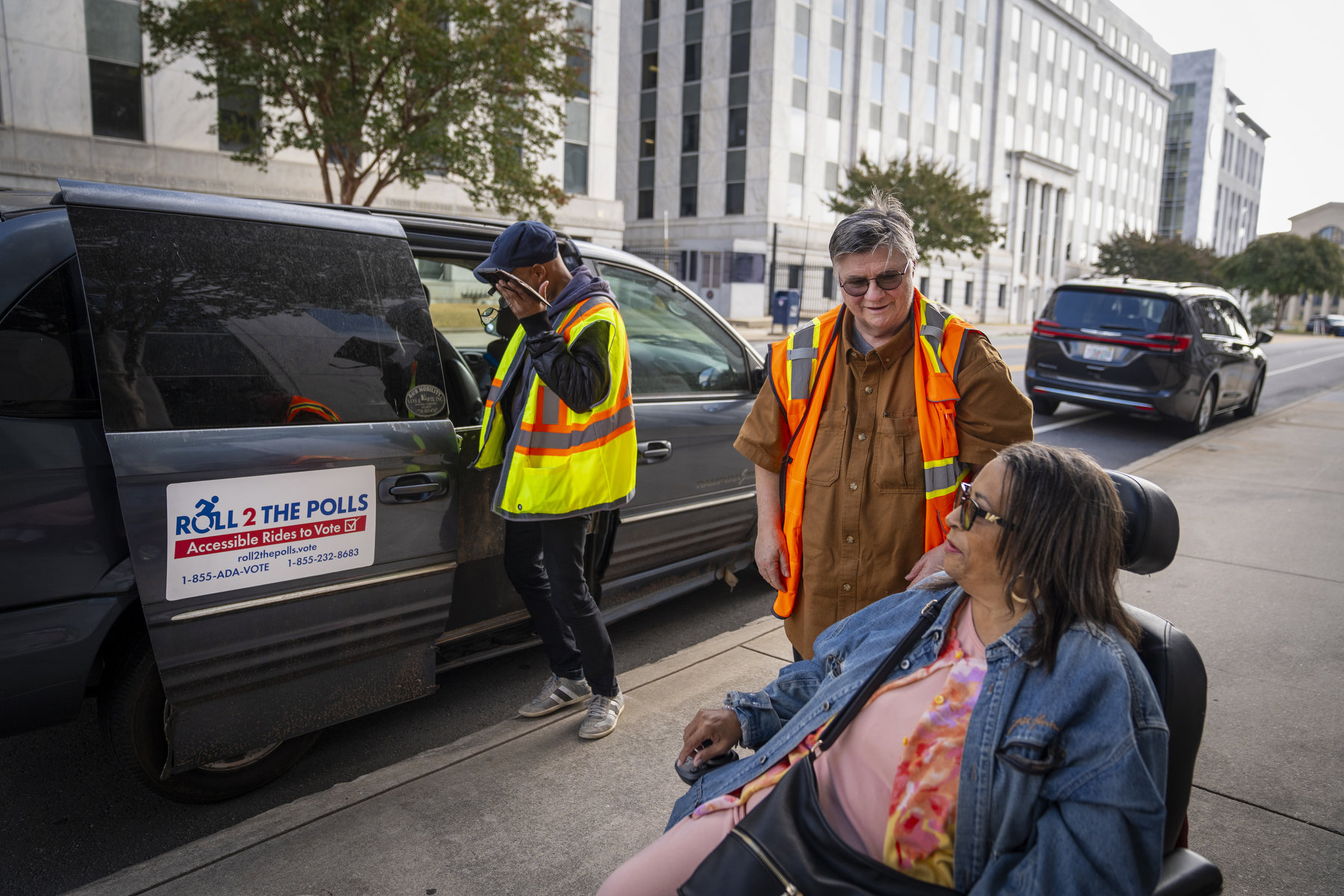 Two people wearing orange safety vests assist a person using a wheelchair on a city street near a van that says "Roll 2 the Polls."