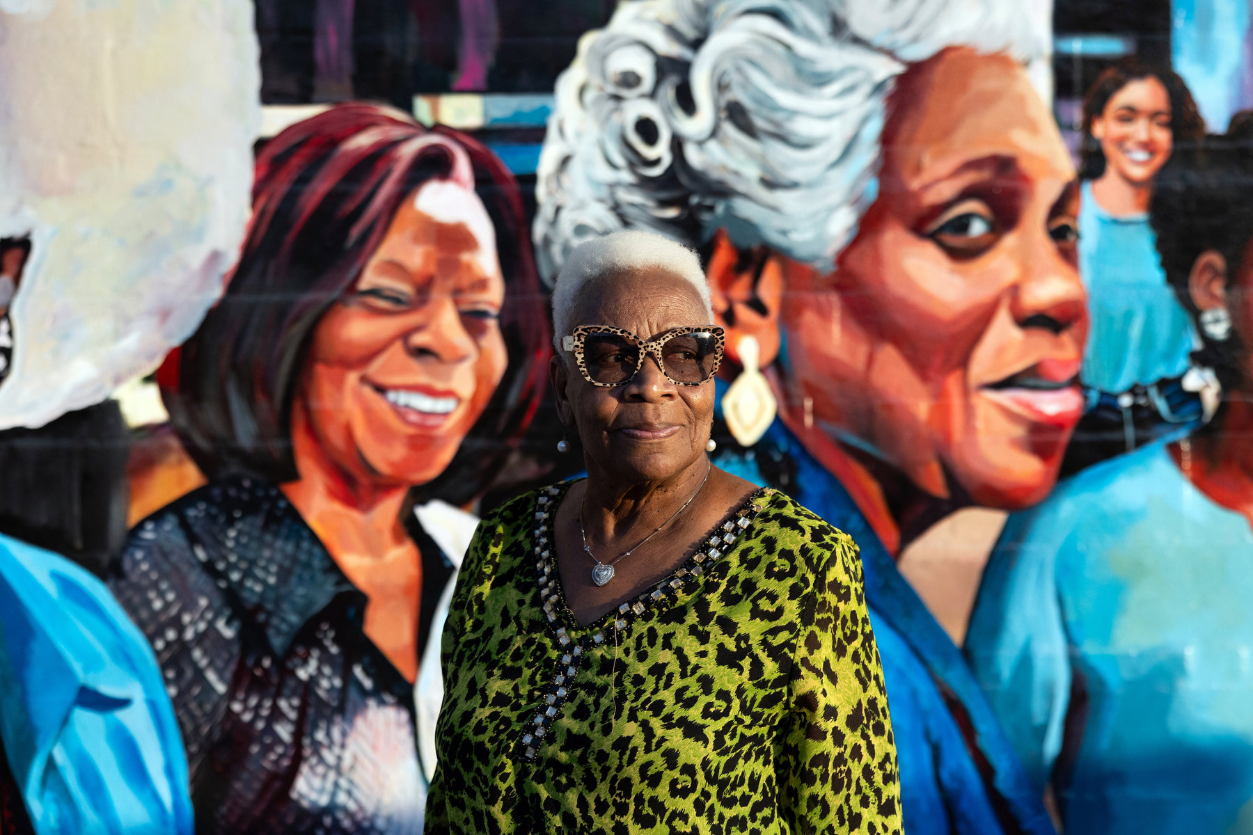 A woman wearing sunglasses stands in front of a mural featuring other Black women.