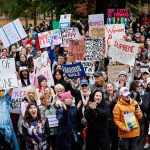 Hundreds gather on Boston Common for a Women's March. People are seen clapping, chanting and holding signs.