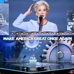 Linda McMahon speaks on stage in front of a screen with gears that says 