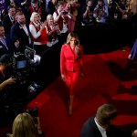 Melania Trump walks on a red carpet, wearing a red suit, surrounded by photographers, camera operators, and supporters holding signs and applauding during the Republican National Convention.