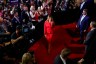 Melania Trump walks on a red carpet, wearing a red suit, surrounded by photographers, camera operators, and supporters holding signs and applauding during the Republican National Convention.