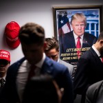 Several men stand in a hallways with a poster of Trump and a MAGA hat hanging on the wall behind them.