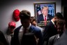 Several men stand in a hallways with a poster of Trump and a MAGA hat hanging on the wall behind them.