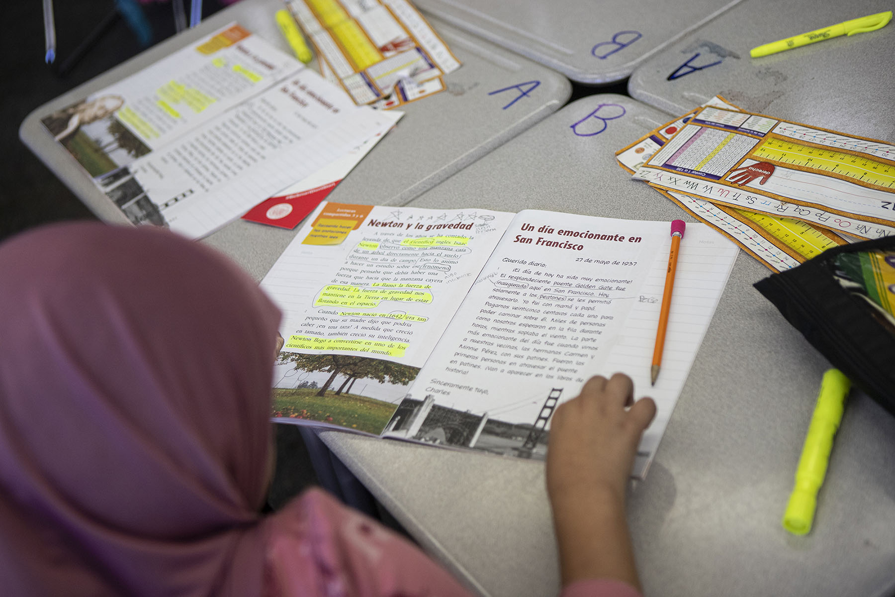 A student sits at her desk and reads her assignment from a Spanish book. Pencils and worksheets are strewn across various desks. The setting is a classroom.