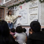 A person in glasses stands and points to a word on a poster board hung on a whiteboard while a group of children sit on the floor and watch.