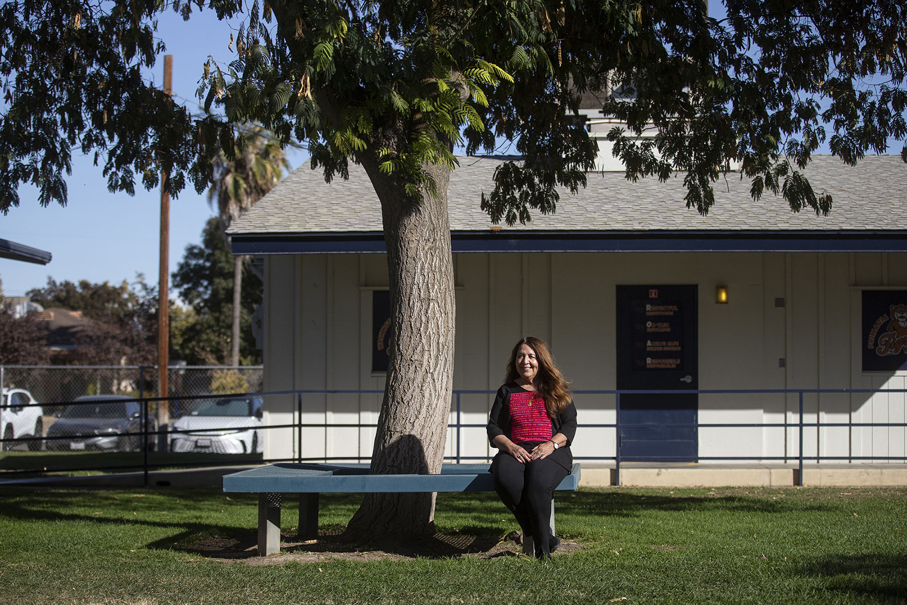 A person sits on a bench underneath a tree in the middle of an elementary school. They're wearing a red and black shirt, with black pants.