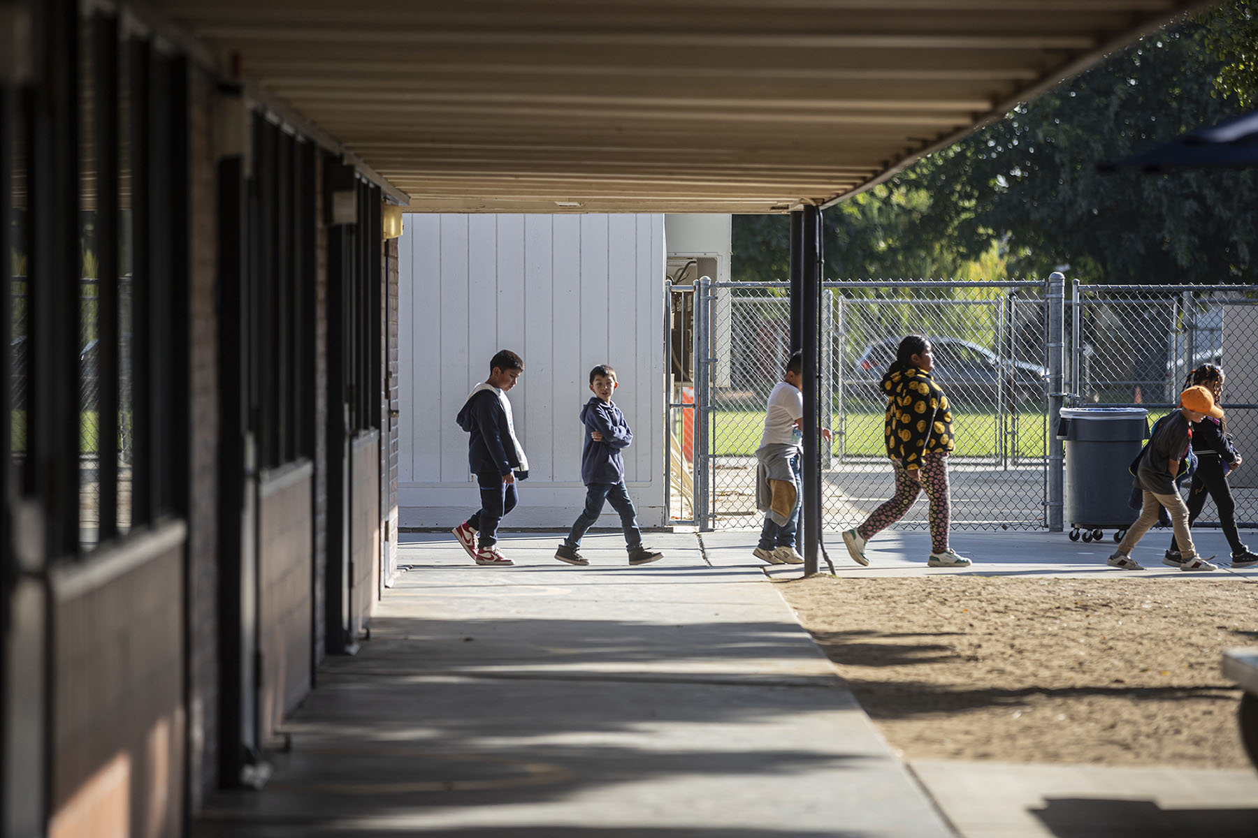 A line of children walk outdoors through a school campus. Fencing and other buildings are visible in the background.