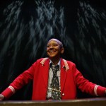 Nikki Giovanni stands at a podium smiling, in a red jacket with lights reflected on the wall behind her.