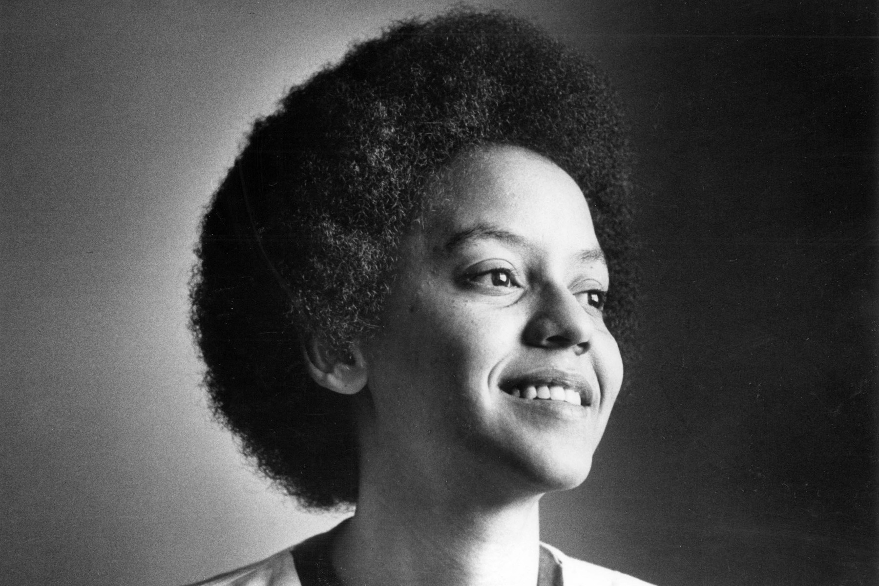 A black and white closeup portrait of a smiling Nikki Giovanni.