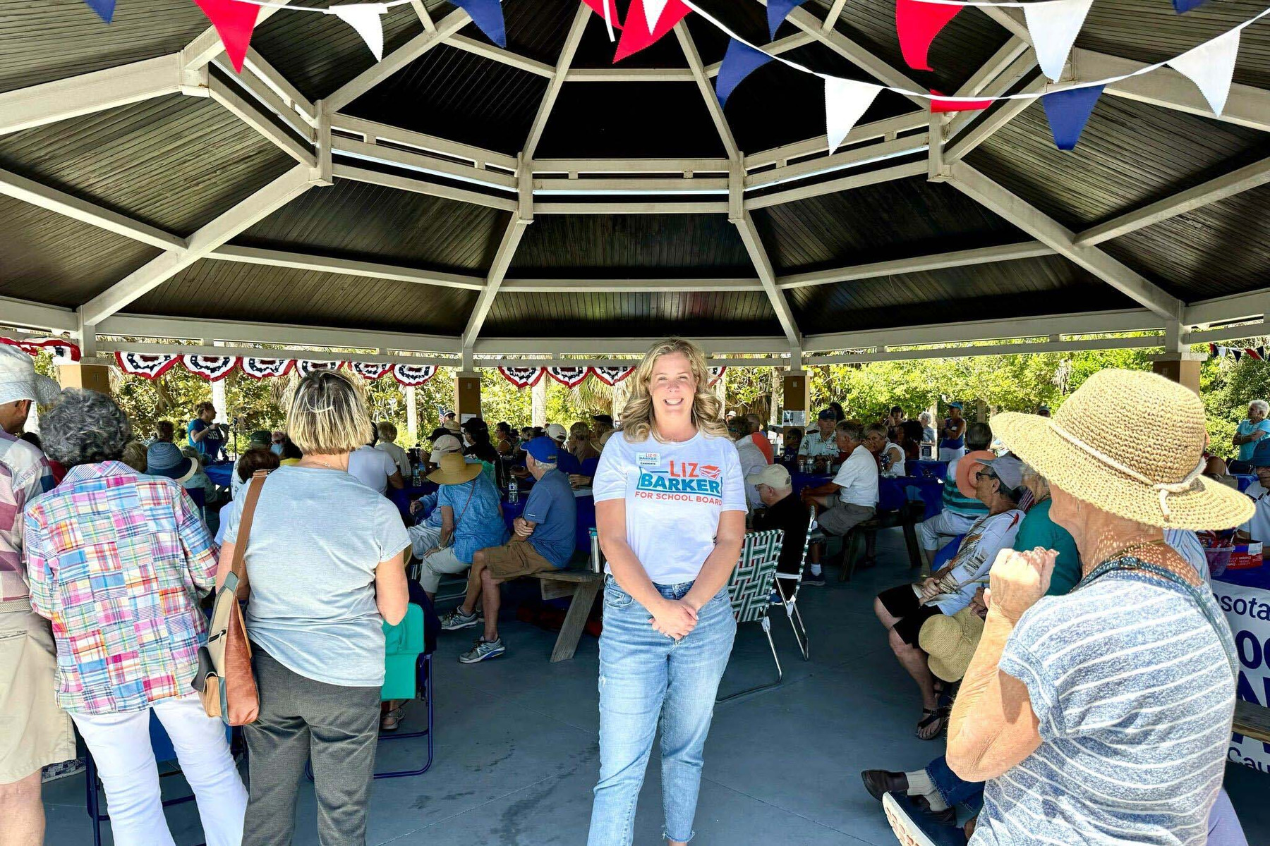 A woman stands in a group of people facing the camera and smiling, wearing a "Liz Barker for School District" shirt