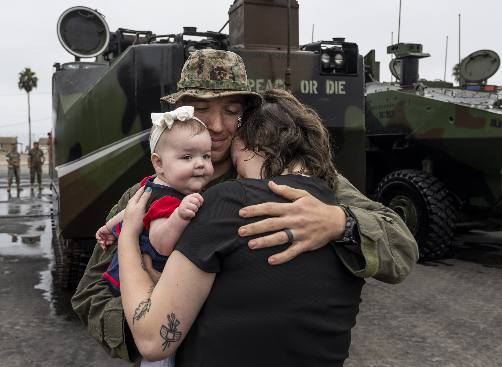 A Marine in uniform hugs his wife and baby in front of a military tank.