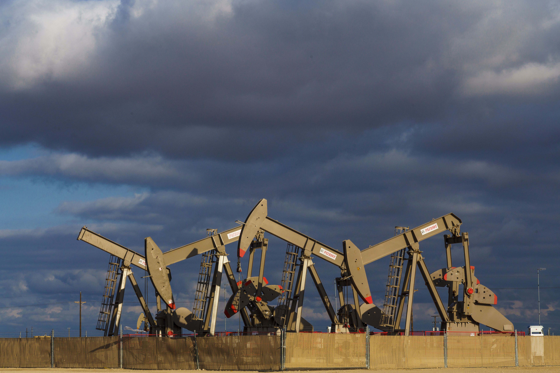 A group of oil wells pump behind a fence against a stormy sky.