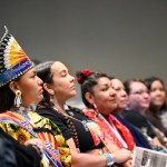 A group of Indigenous women sit in a line, listening to an unseen speaker.