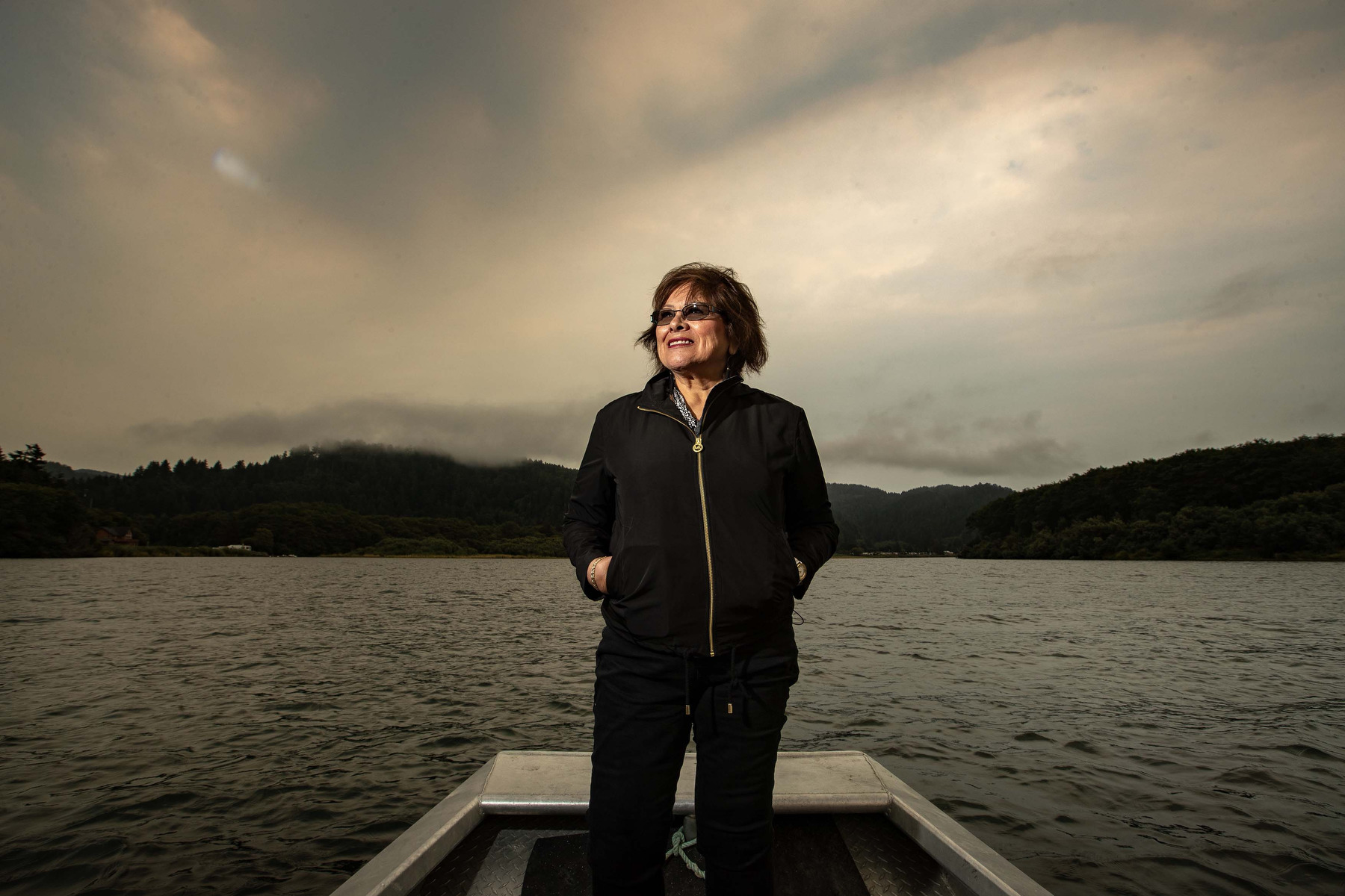 A woman stands on a boat in a lake with mountains and clouds behind her.