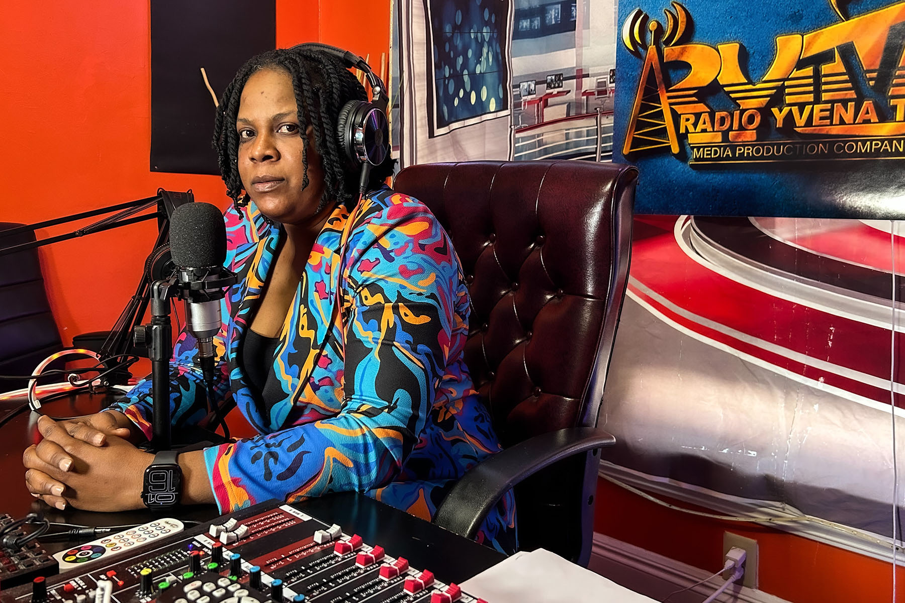Yvena Jean François sits at her desk in her podcasting studio on Inauguration Day, January 20, 2024, in Springfield, Ohio. She wears a colorful patterned jacket and headphones while seated in front of a microphone and soundboard, with a vibrant studio backdrop featuring the logo of Radio Yvena TV.