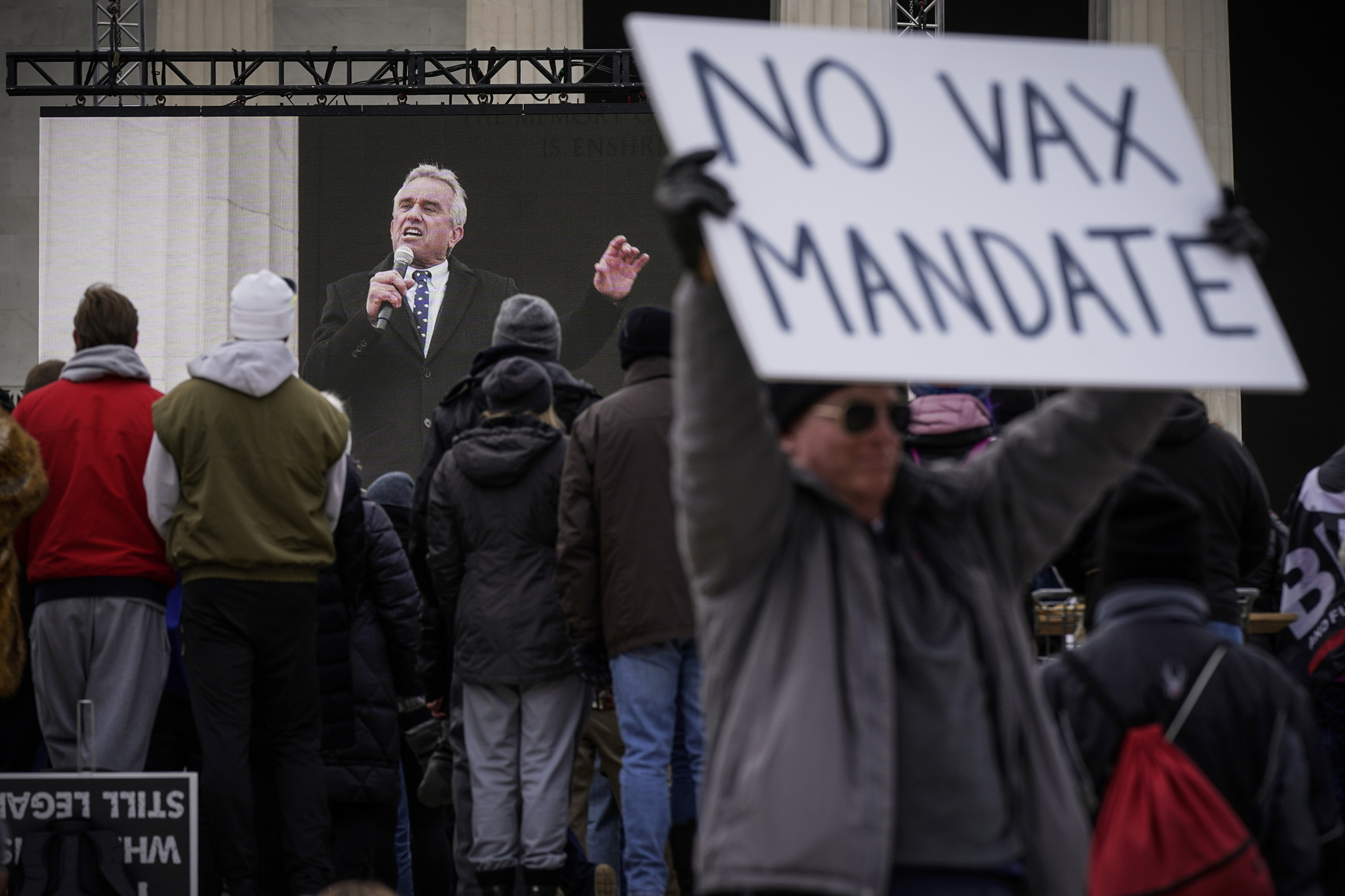 A man holds a sign that says "no vax mandate" while Kennedy speaks onstage in the background.