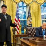 Elon Musk, wearing a black Make America Great Again cap, stands in the Oval Office next to President Donald Trump, who is seated at the Resolute Desk