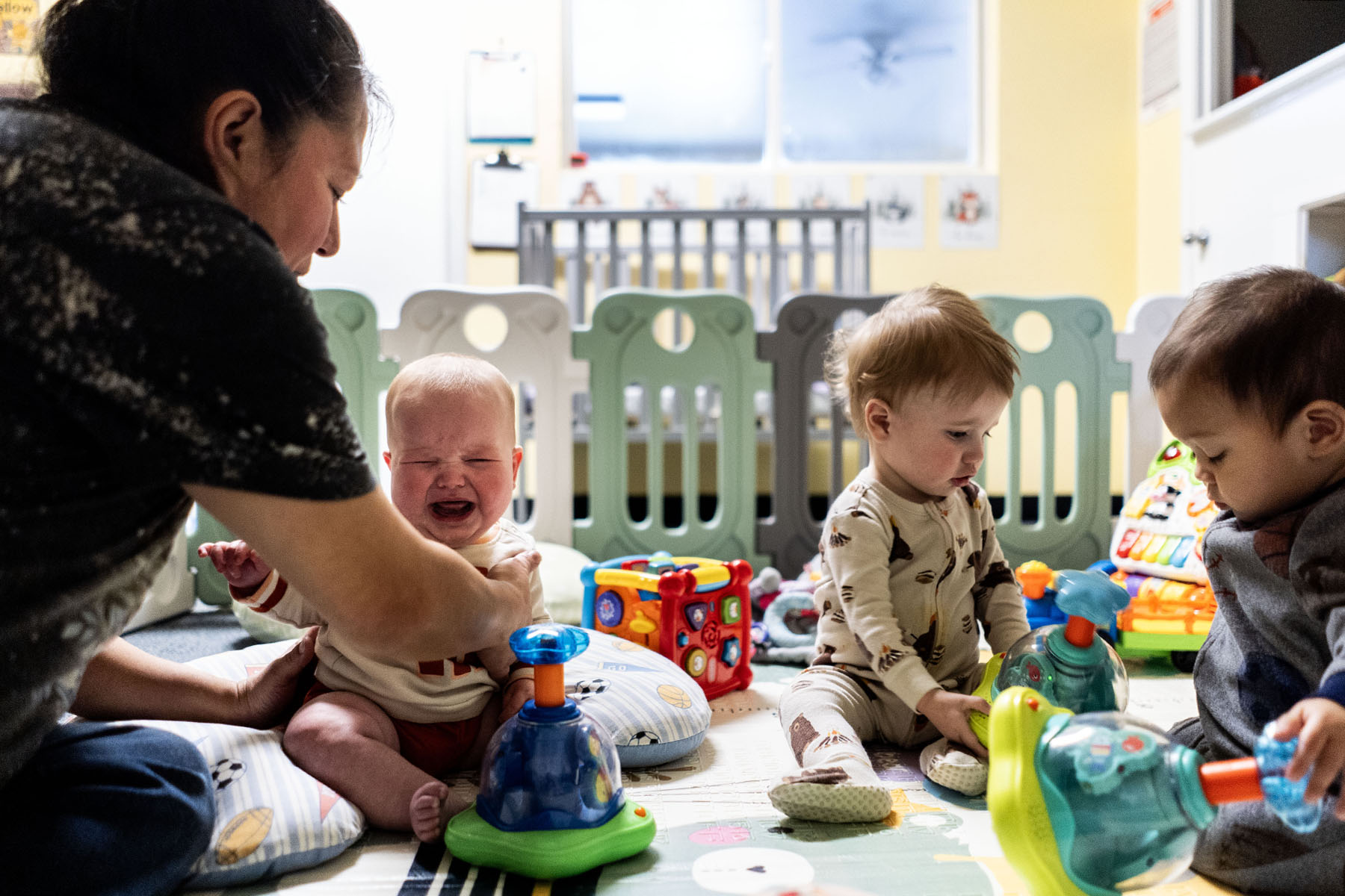 A teacher comforts a young child at a daycare as other toddlers play.