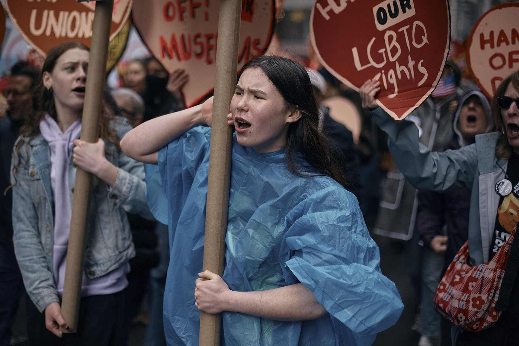 A young woman in a blue rain poncho shouts during a protest in New York City. She holds a signpost and is surrounded by other demonstrators holding red heart-shaped signs that read slogans like “Hands Off Our LGBTQ Rights.”