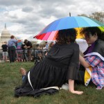 Two people sit on the grass under a colorful umbrella.