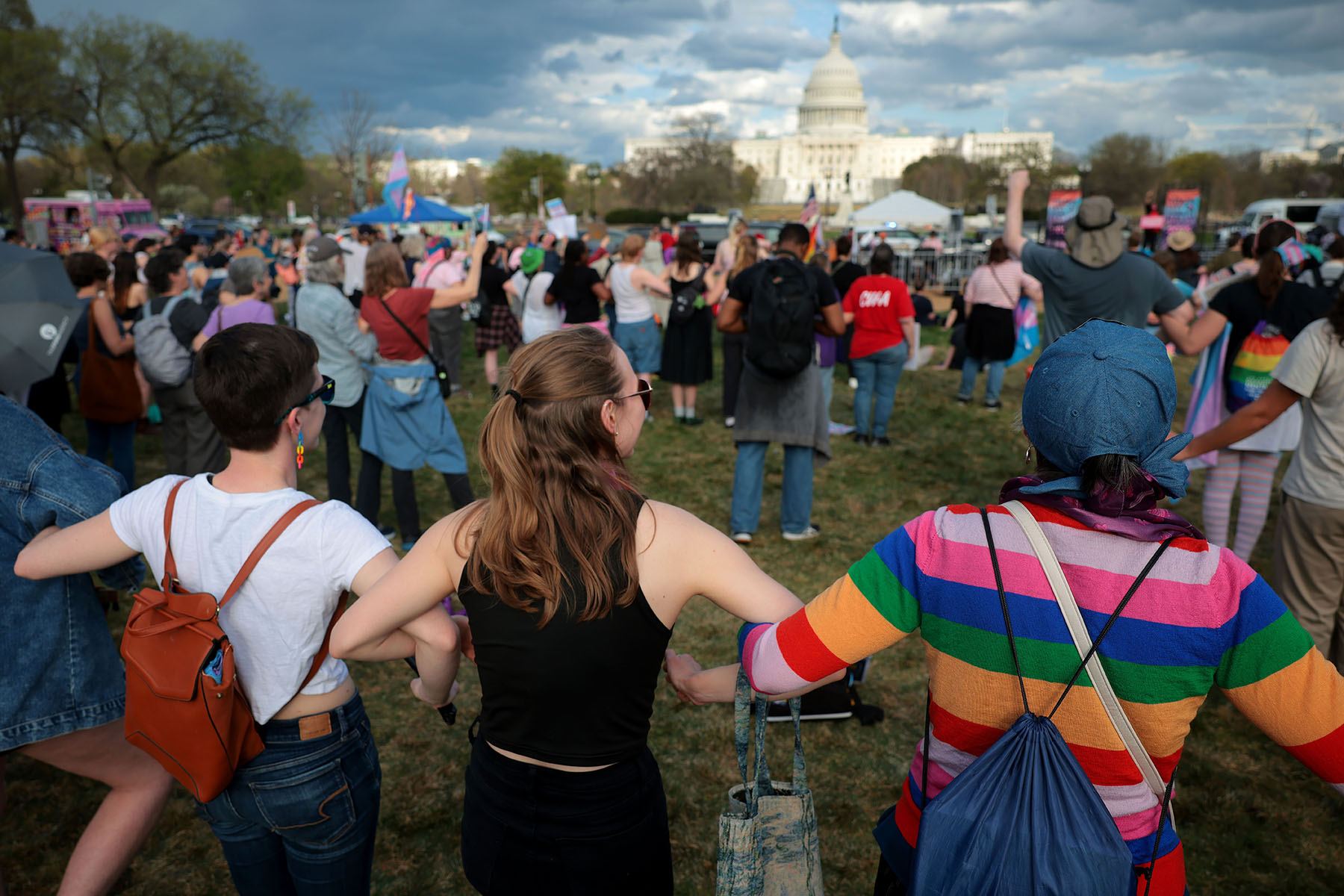 A crowd of demonstrators, many wearing or carrying Pride flags and gear, join hands in a line facing the Capitol during a rally in Washington, D.C.